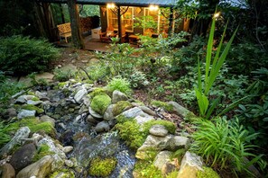 Uma cabana rústica com um deck de madeira, uma pequena cascata e um jardim com diversas plantas.