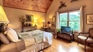A cozy room with a bed, a TV, a wooden ceiling, and a window with a view of greenery. - Scenic Mountain Cabin with Hot Tub, Game Room & Firepit near Silver Run Falls – Cashiers, NC (Cashiers)
