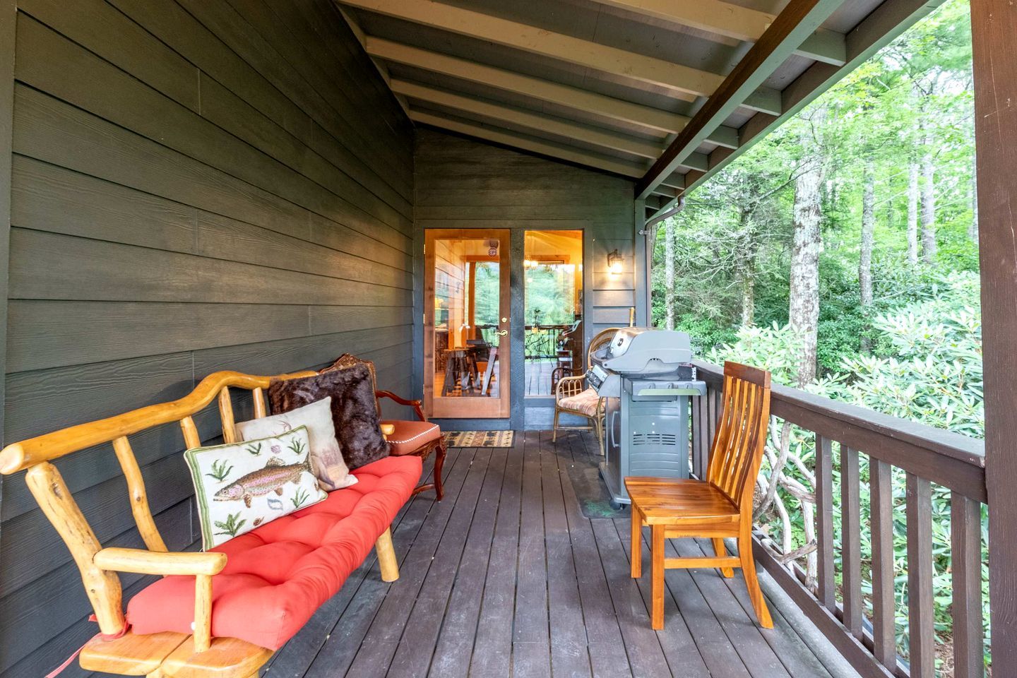 A covered porch with a wooden bench and a grill, overlooking a forested area.