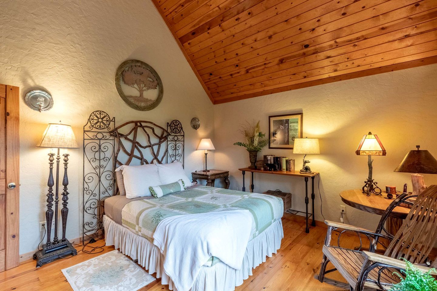 A bedroom with a wooden ceiling, a bed with a decorative headboard, two bedside tables with lamps, a small wooden side table, a chair, and a framed picture on the wall.