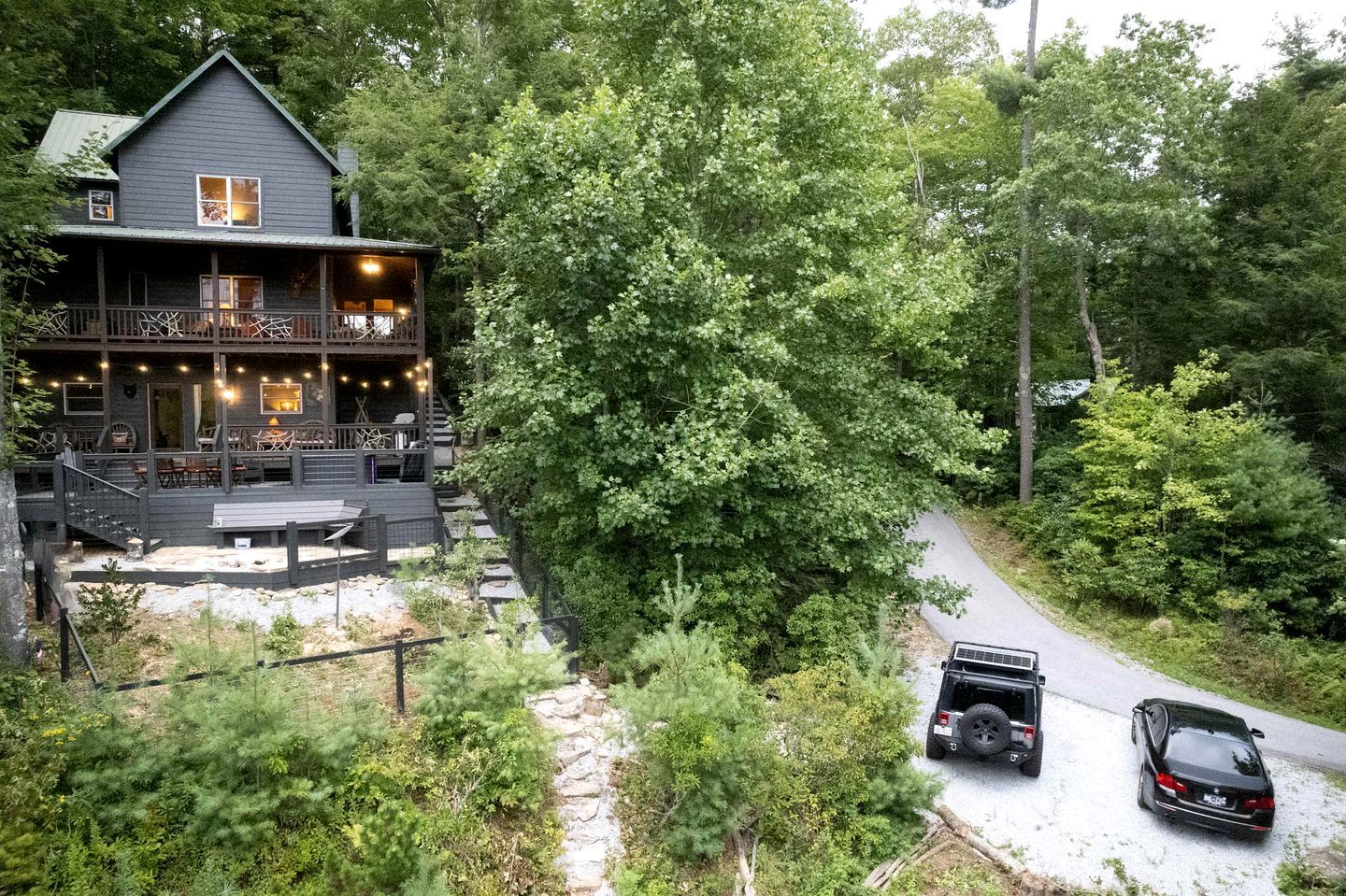 A cabin with a deck, surrounded by dense forest, and two vehicles parked on a gravel path.