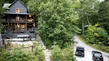 A cabin with a deck, surrounded by dense forest, and two vehicles parked on a gravel path.