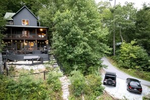 Una cabaña con terraza, rodeada de un bosque frondoso, y dos vehículos estacionados en un camino de grava.