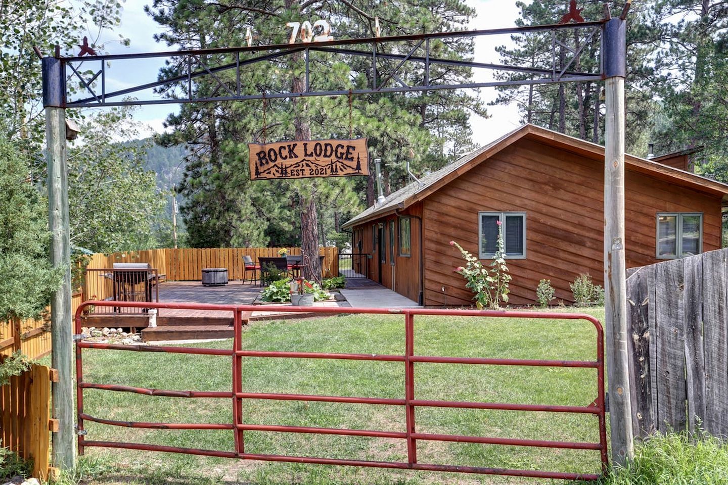 A wooden cabin with a porch, a fenced yard, and a sign indicating 'Rock Lodge' with a date of establishment.