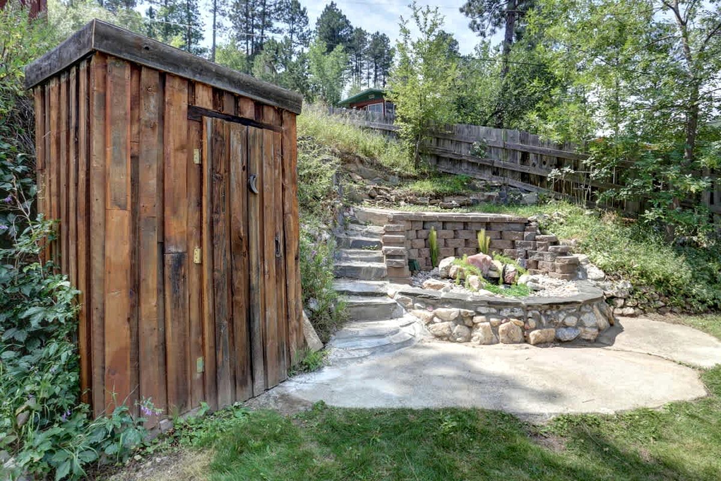 Ein Holler Schuppen, eine Feuerstelle aus Stein und ein Betonweg in einem Garten.