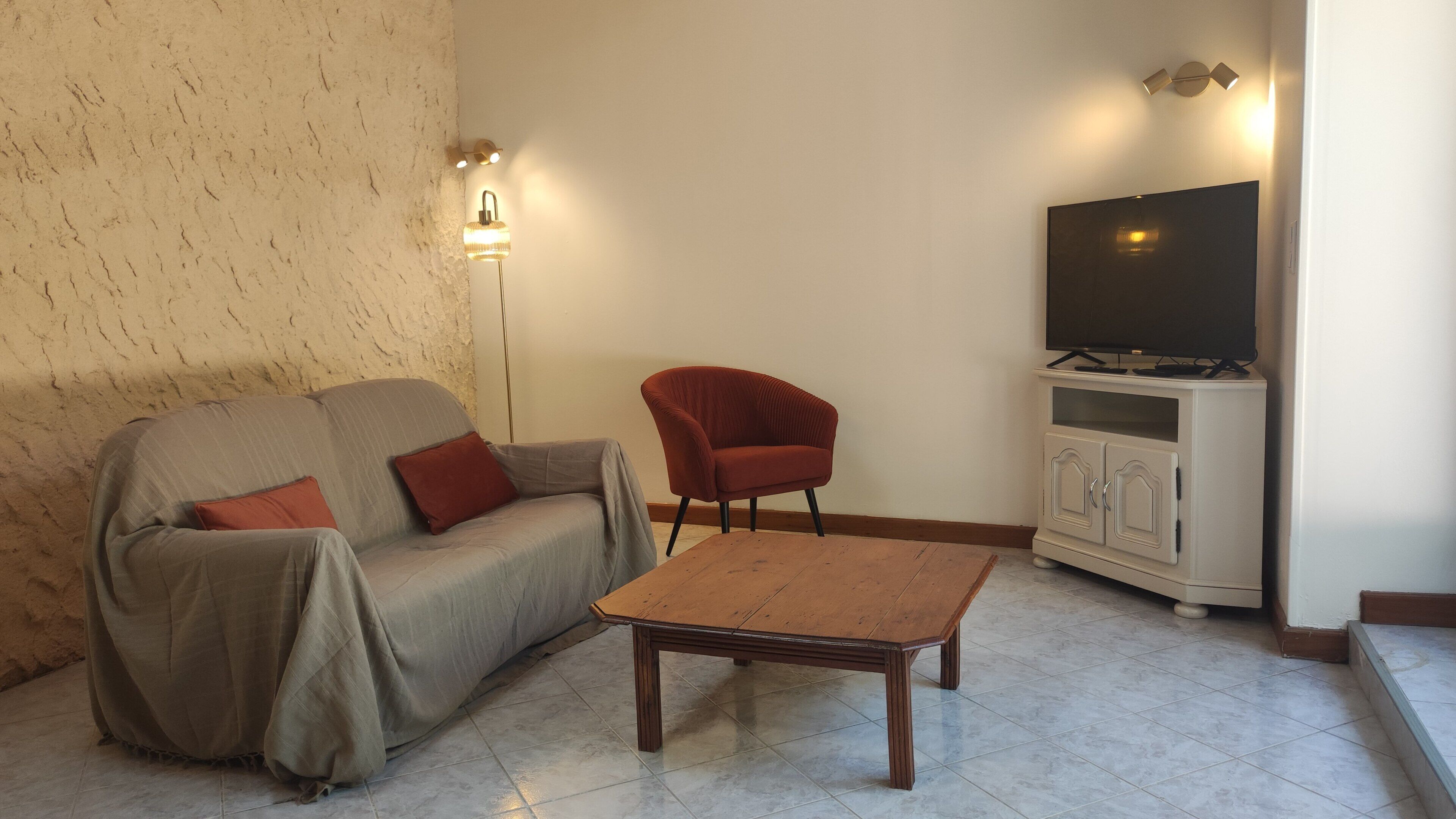 Uma sala de estar com sofá, uma poltrona vermelha, mesa de centro, televisão sobre um móvel e luminárias fixadas na parede.