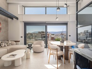 A modern living room with a large sliding glass door leading to a balcony, offering a city view. - Cosmos Penthouse Suite (Chania)