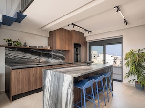 A modern kitchen with a marble island, wooden cabinets, and blue bar stools. - Cosmos Penthouse Suite (Chania)