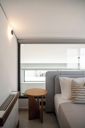 A modern hotel room with a bed, a small round table, a window with a view, and a wall-mounted light fixture. - Cosmos Penthouse Suite (Chania)