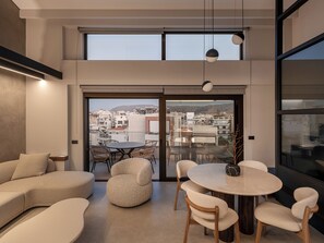 Un salón moderno con una mesa redonda y sillas, un sofá y vistas a un paisaje urbano a través de ventanas amplias.