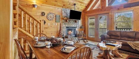 Interior de una cabaña de madera con una mesa de comedor, chimenea, televisión y un rincón de estar acogedor.