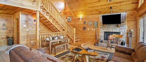 Interior de una cabaña de troncos con escalera de madera, chimenea, televisor de pantalla plana y una zona de estar acogedora.