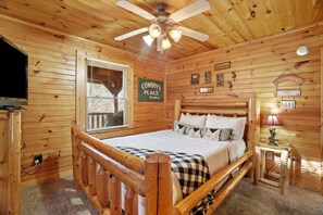 A wooden cabin bedroom with a bed, a ceiling fan, a TV, and a window with blinds. - Stunning Log Cabin with Movie Theatre & Hot Tub near Arts and Crafts District in Gatlinburg, TN (Gatlinburg)