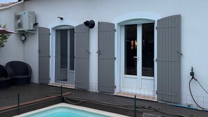 Una piscina con borde de baldosas, un edificio blanco con contraventanas grises y un equipo de aire acondicionado.
