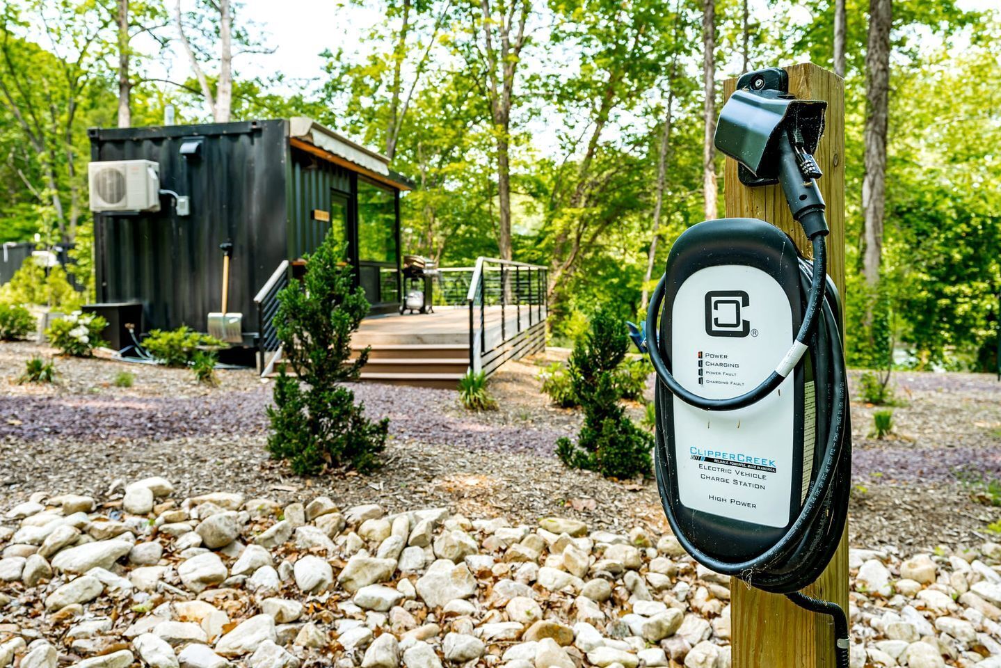 Una estación de carga para vehículos eléctricos con un poste de madera, un camino de grava y una cabaña pequeña al fondo.