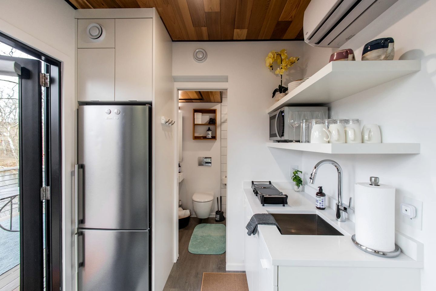 A compact kitchen with a stainless steel refrigerator, a sink with a faucet, and a countertop with a microwave and a small plant.
