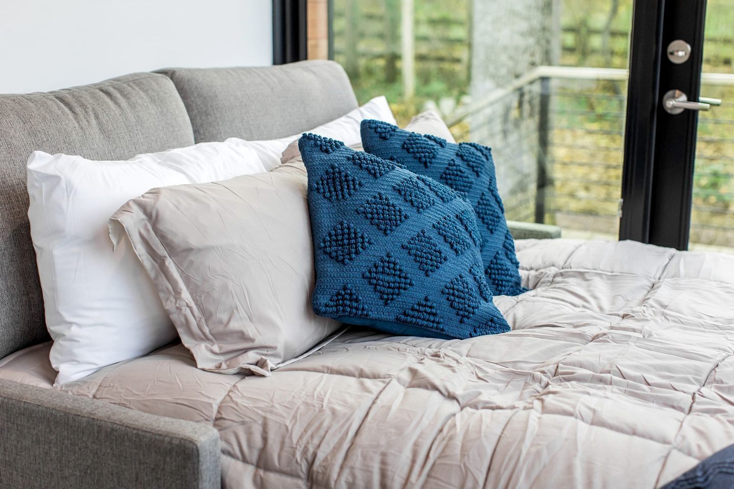 Una cama con edredón color beige, dos almohadas y una manta tejida azul.