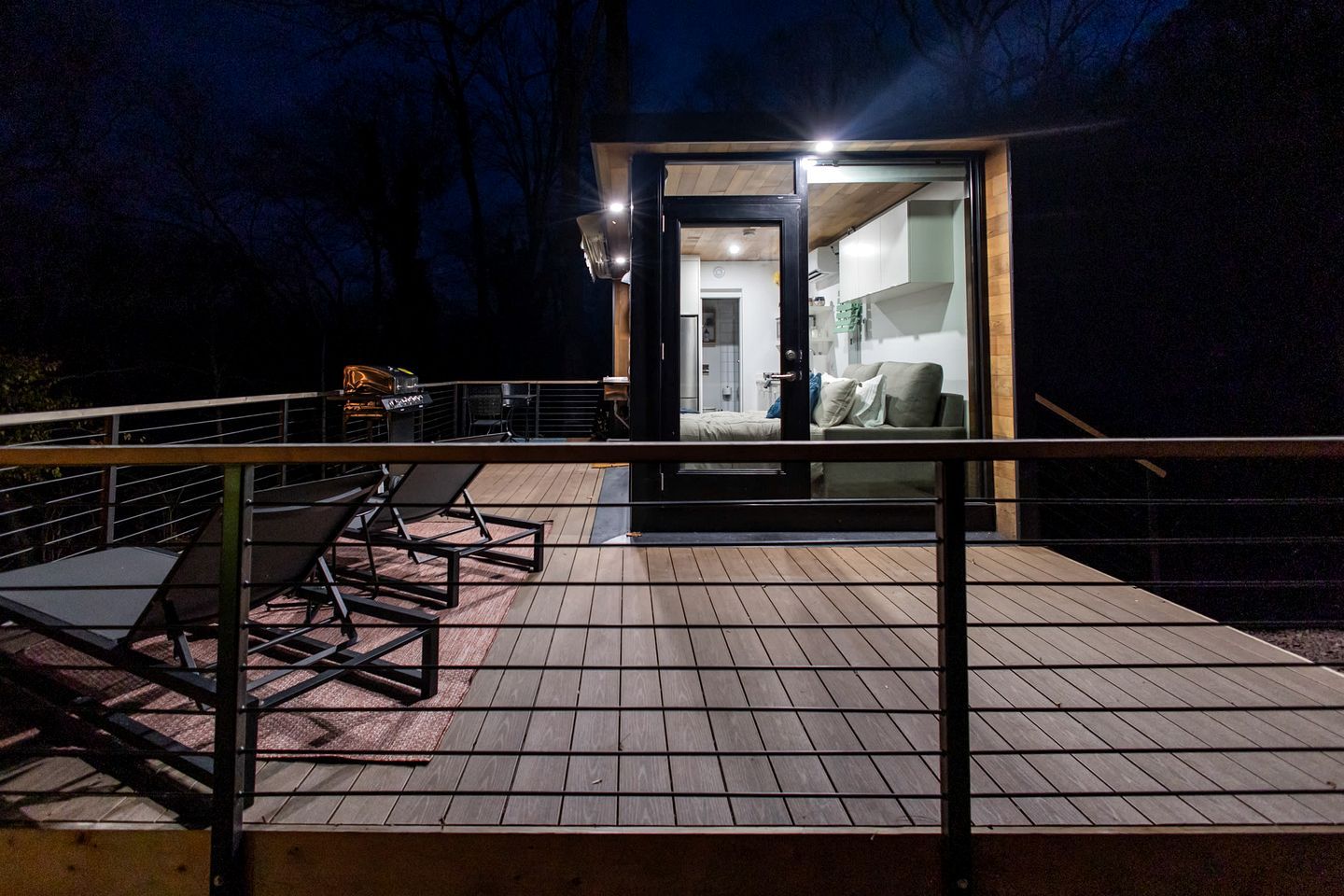 A wooden deck with outdoor furniture, a glass door leading to a room with a bed and a sofa, and a staircase.