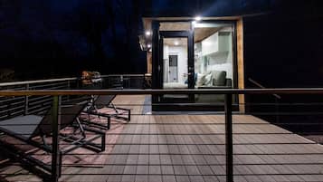 A wooden deck with outdoor furniture, a glass door leading to a room with a bed and a sofa, and a staircase.