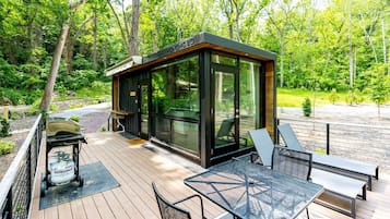 Un'area salotto esterna moderna con struttura in vetro, terrazza in legno e mobili in metallo.