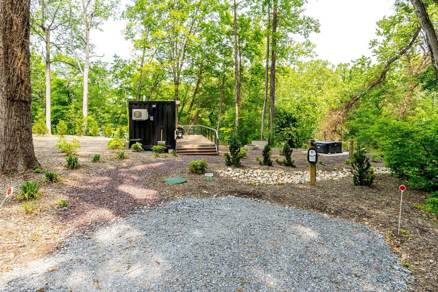 A small cabin with a gravel driveway and a wooden deck, surrounded by trees and a gravel path.