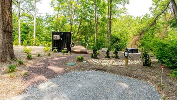 A small cabin with a gravel driveway and a wooden deck, surrounded by trees and a gravel path.