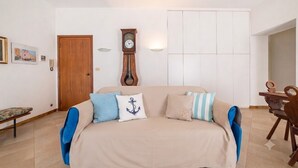 A living room with a sofa, a wooden door, a wall-mounted clock, and a dining area with a wooden table and chairs. - Appartamento il Corsaro (Imperia)
