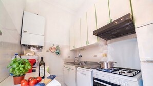 A compact kitchen with white cabinets, a gas stove, a built-in oven, and a small countertop with a plant, tomatoes, and a bottle. - Appartamento il Corsaro (Imperia)