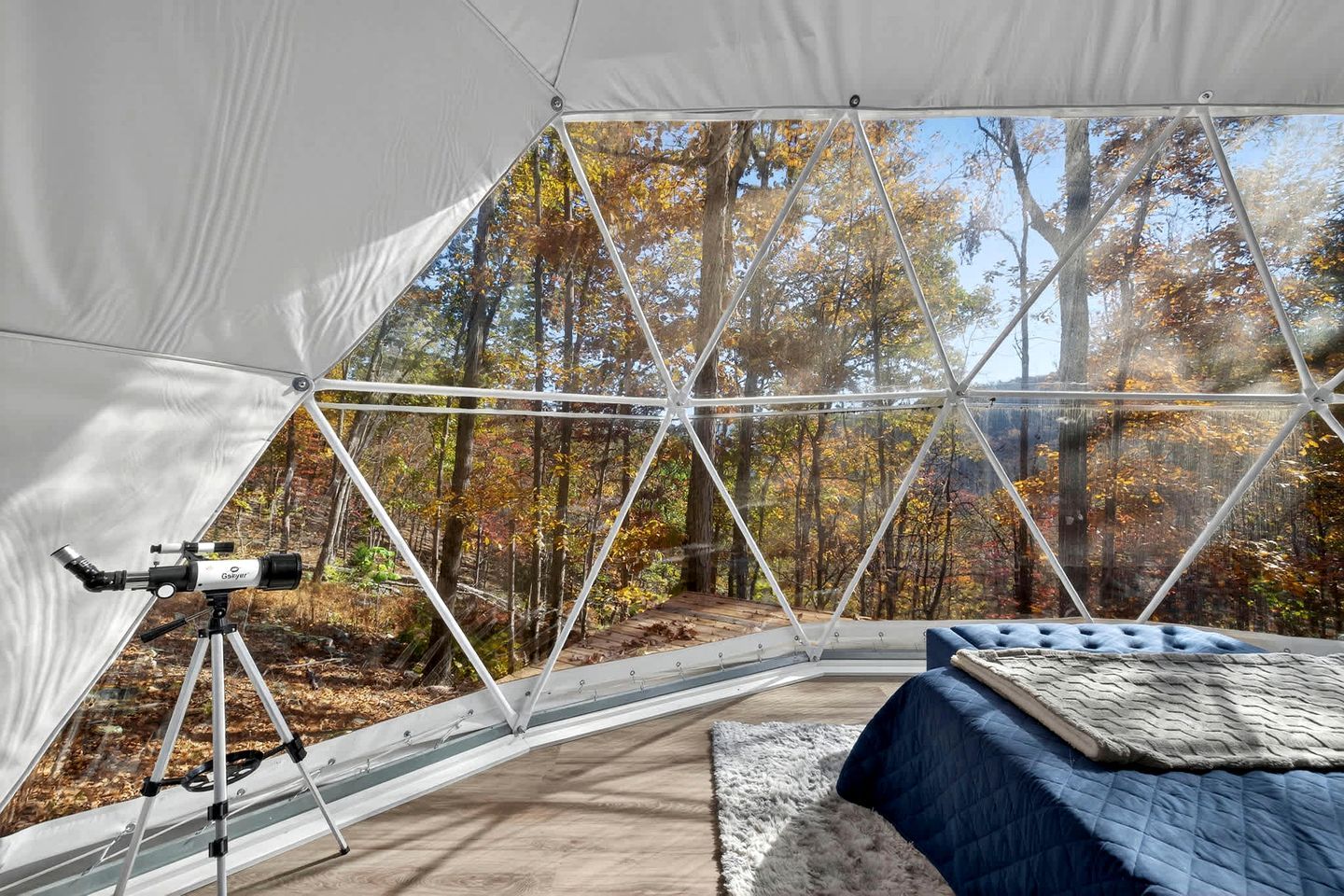A geodesic dome tent with a telescope, a bed, and a view of a forest.