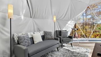 A tented room with a grey sofa, a chair, a telescope, and a view of autumn trees.