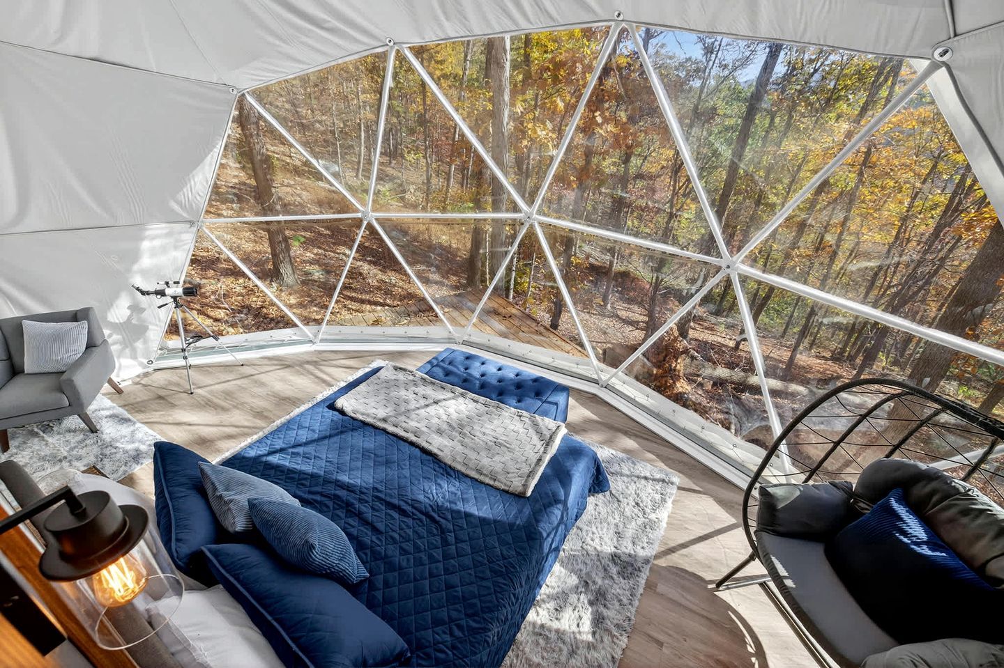 A tented room with a bed, a chair, and a view of a forest.