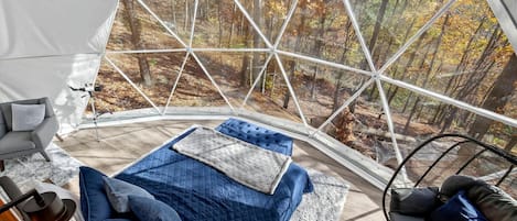 A tented room with a bed, a chair, and a view of a forest.