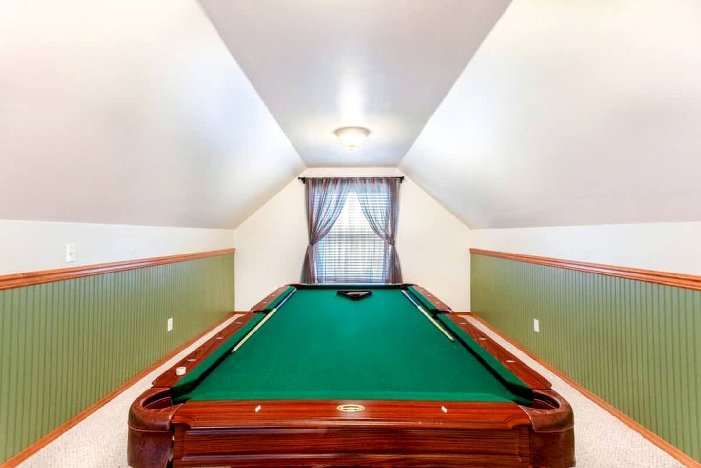 A pool table in a room with green paneling, a window with curtains, and a sloped ceiling.