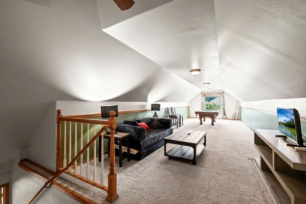 Un loft spacieux avec un plafond haut, un escalier en bois, un canapé, une table basse, une table de billard et un téléviseur.