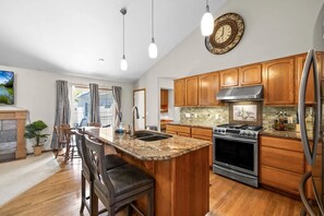 A modern kitchen with wooden cabinets, a granite countertop, stainless steel appliances, and a large island with bar stools. - Spacious Retreat with Fireplace, Game Room & Fire Pit in Post Falls, Idaho (Post Falls)