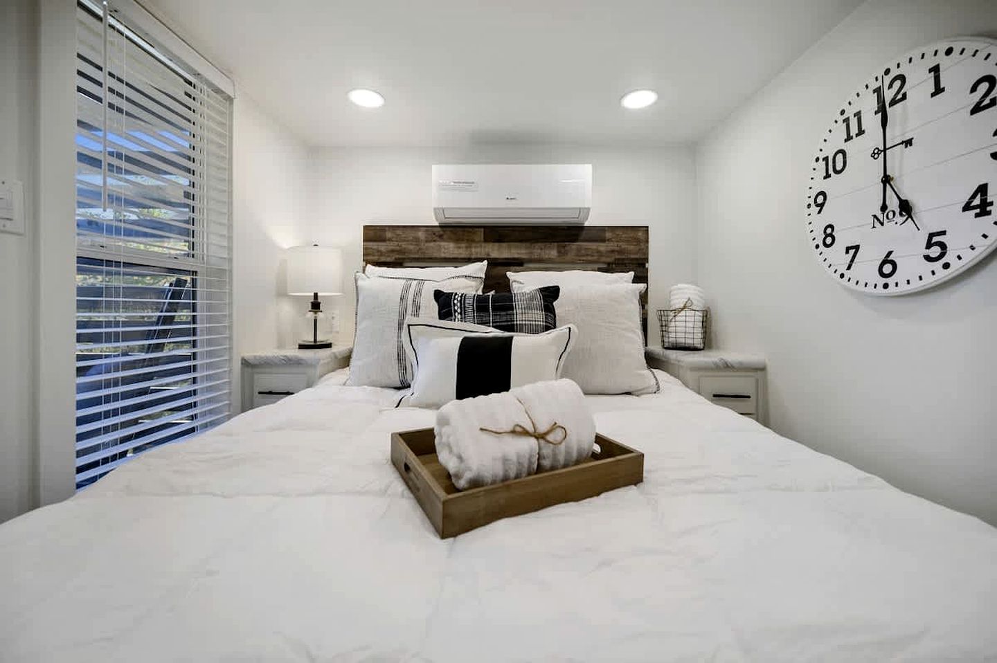 A bedroom with a bed, a bedside table, a lamp, a clock, and a window with blinds.