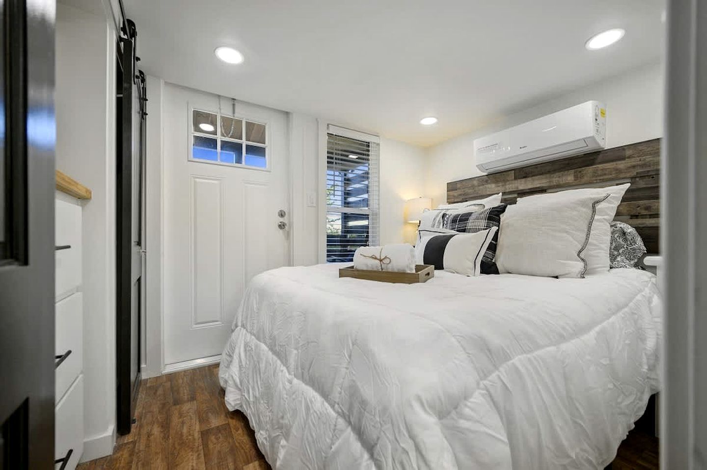 A bedroom with a bed, a bedside table, a window with blinds, an air conditioning unit, and a door.