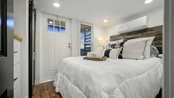 A bedroom with a bed, a bedside table, a window with blinds, an air conditioning unit, and a door.