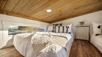 A bedroom with a wooden ceiling, a large bed with white bedding, a bedside table with a plant, and a window with a view of trees.