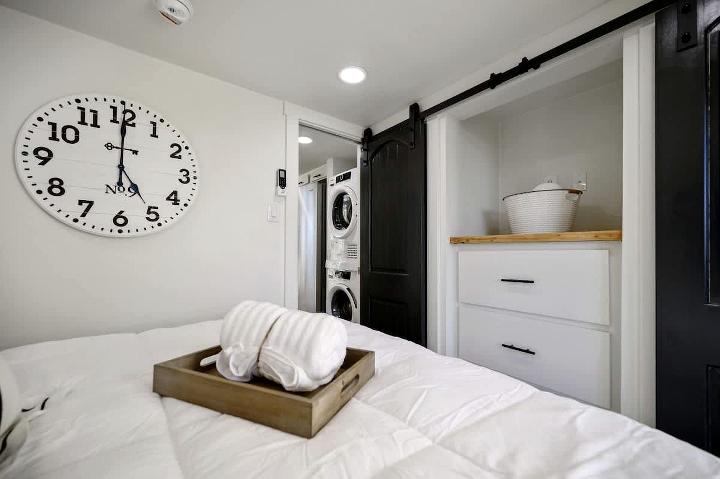 Una camera d'albergo con un letto, una sveglia, una lavatrice e una mensola in legno con un cesto.