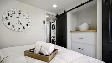 A hotel room with a bed, a clock, a washing machine, and a wooden shelf with a basket.