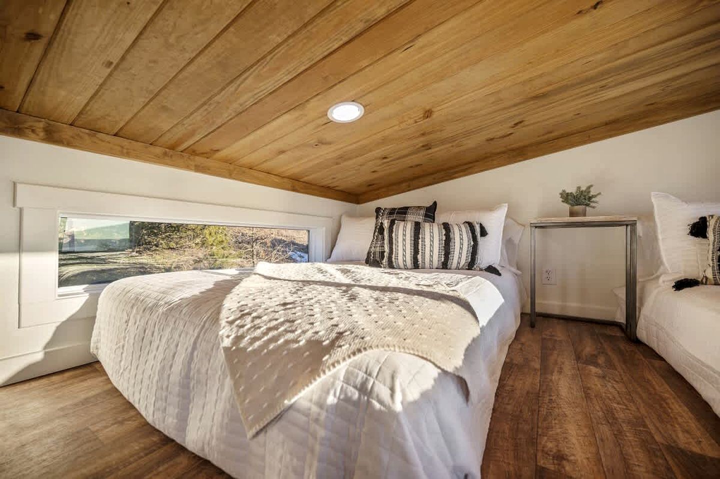 Un dormitorio con techo de madera, una cama grande con cubrecama blanco, una mesita de noche con una planta y una ventana con vista a árboles.