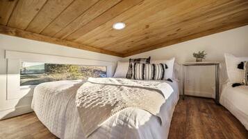 Un dormitorio con techo de madera, una cama grande con cubrecama blanco, una mesita de noche con una planta y una ventana con vista a árboles.