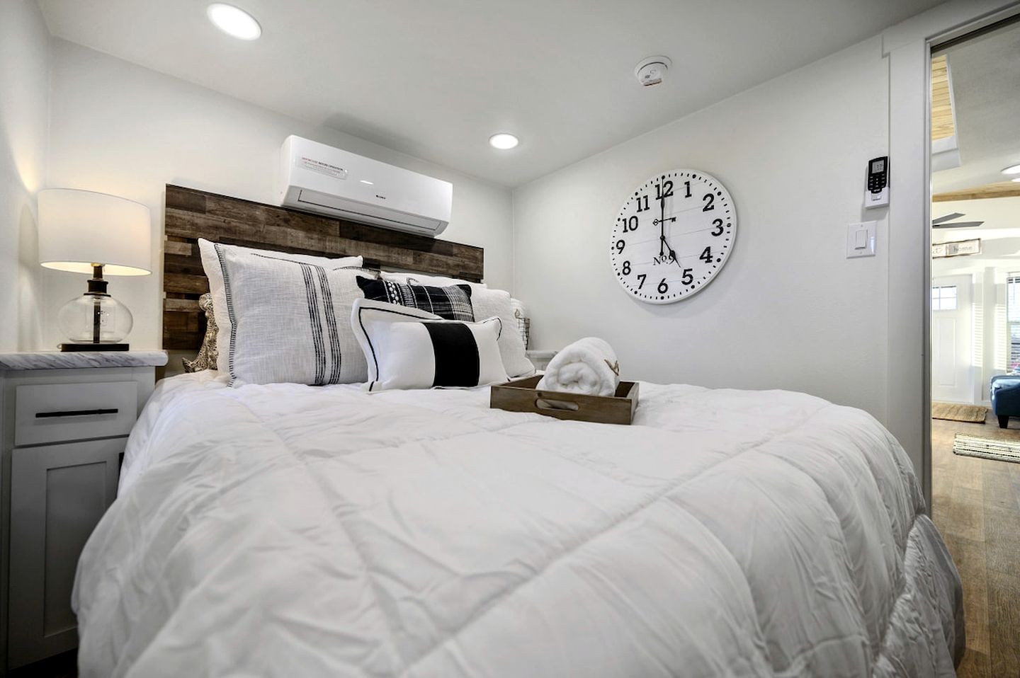 A hotel room with a large bed, a wall clock, an air conditioning unit, and a bedside table with a lamp.