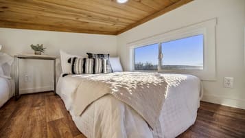 A bedroom with a wooden ceiling, a large window, a bed with a white bedspread, a bedside table with a plant, and a nightstand.