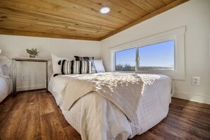 A bedroom with a wooden ceiling, a large window, a bed with a white bedspread, a bedside table with a plant, and a nightstand.