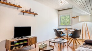 A cozy living room with a flat-screen TV, wooden furniture, a dining area with a table and chairs, and a window with a view of trees.