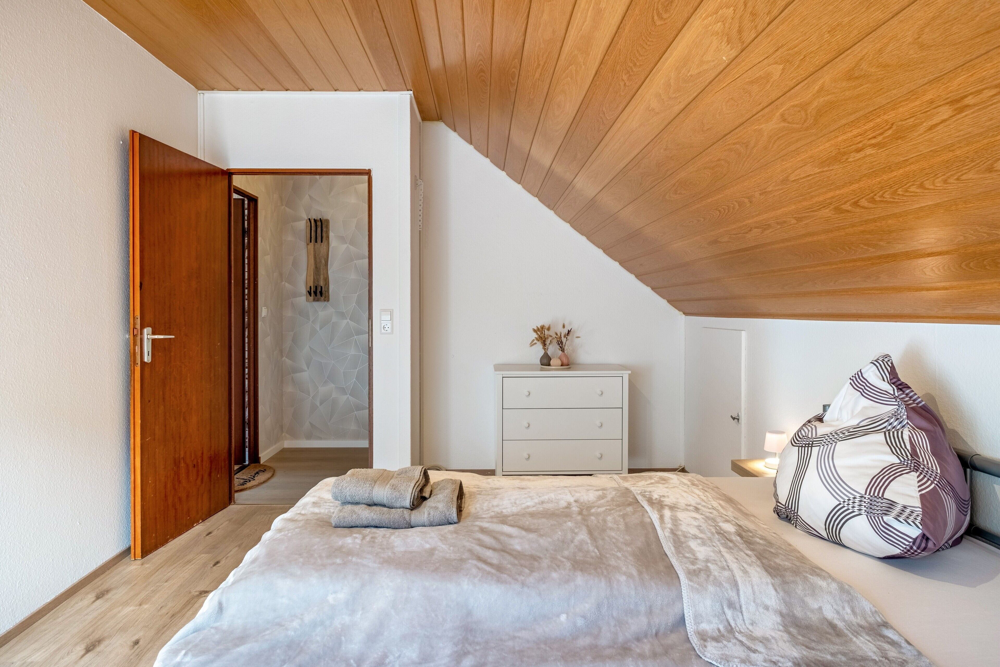 Ein Schlafzimmer mit geneigter Decke, einem Bett mit weißer Bettdecke, einer Holztür, einer weißen Kommode und einem Nachttisch mit Lampe.