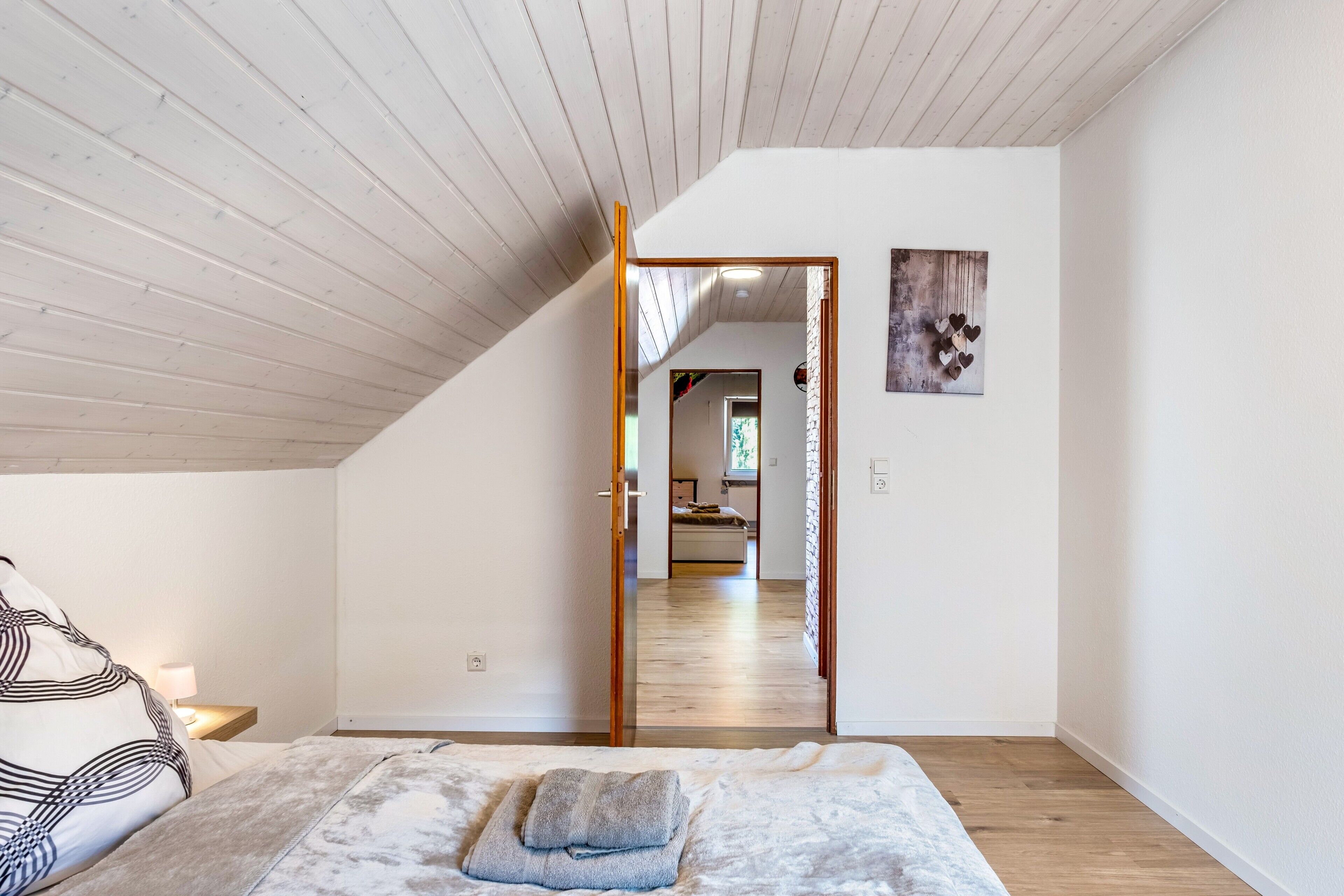 A bedroom with a bed, a nightstand, a lamp, a wooden ceiling, and a door leading to another room.