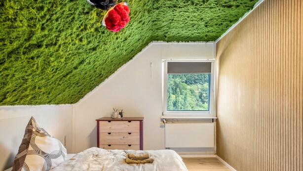 A bedroom with a sloped ceiling covered in green moss, a bed with white bedding, a nightstand with a lamp, and a window with a view of trees.